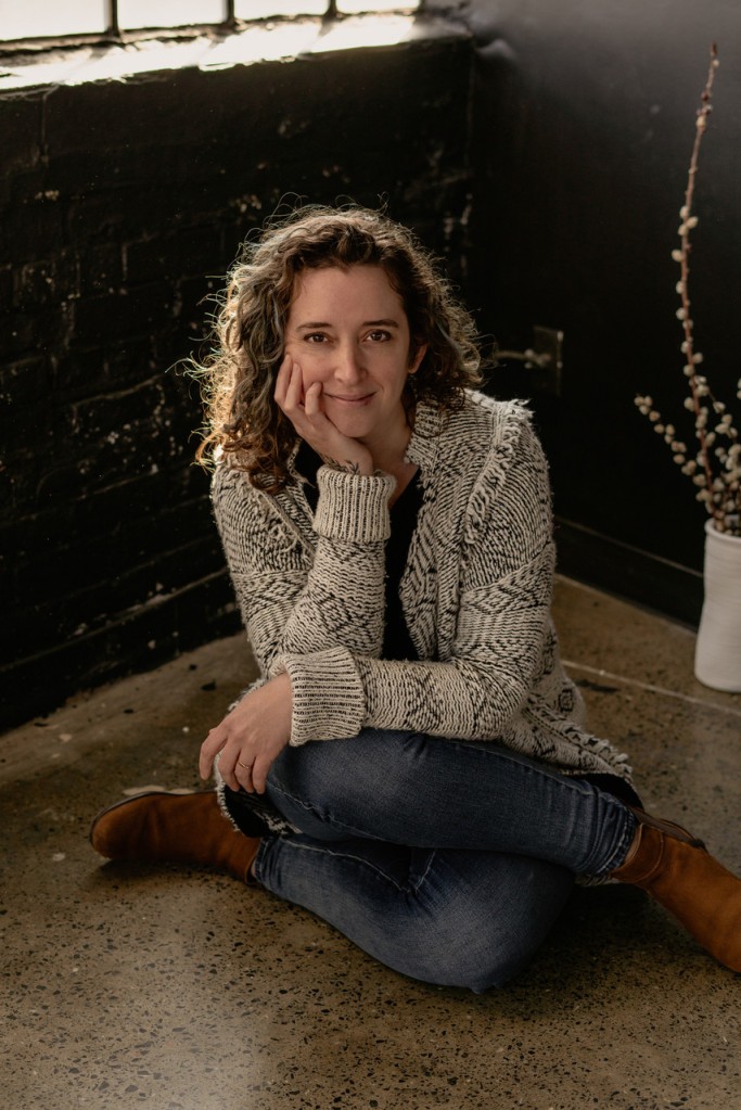 a photo of Jenn seated cross-legged on the ground smiling at the camera; wearing a black and white patterned sweater and jeans